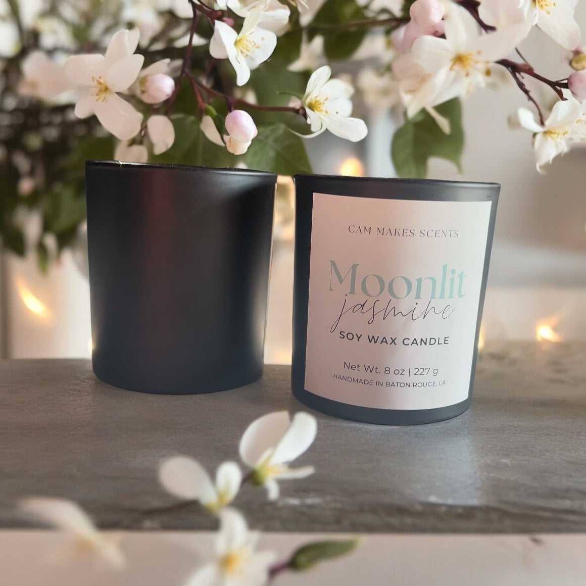 8 oz soy wax candle in a black jar labeled Moonlit jasmine and is surrounded by white jasmine flowers sitting on concrete table