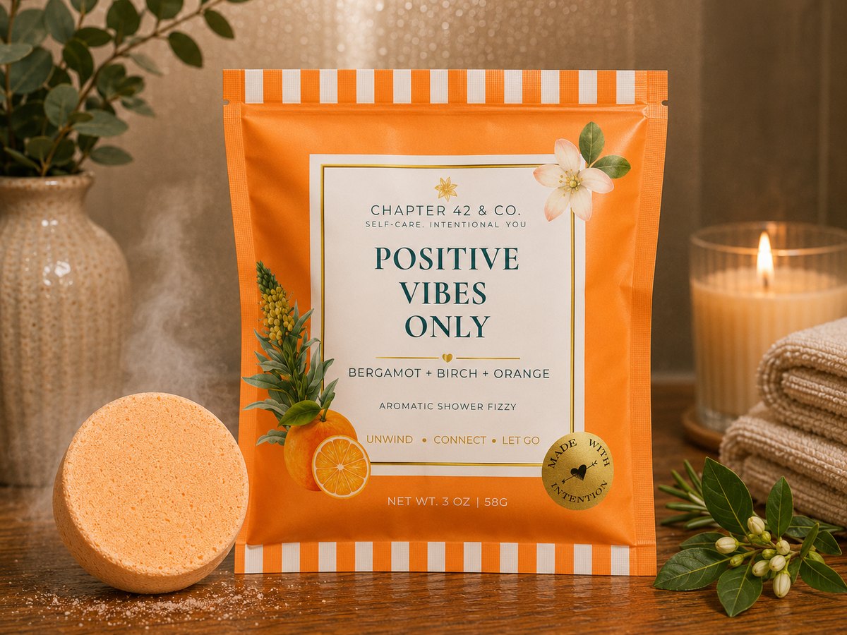 A stand-up pouch of Chapter 42 & Co. Positive Vibes Only aromatic shower fizzy in Bergamot, Birch, and Orange scent, displayed on a wooden surface with a round bath bomb and spa props including a candle and folded towels.
