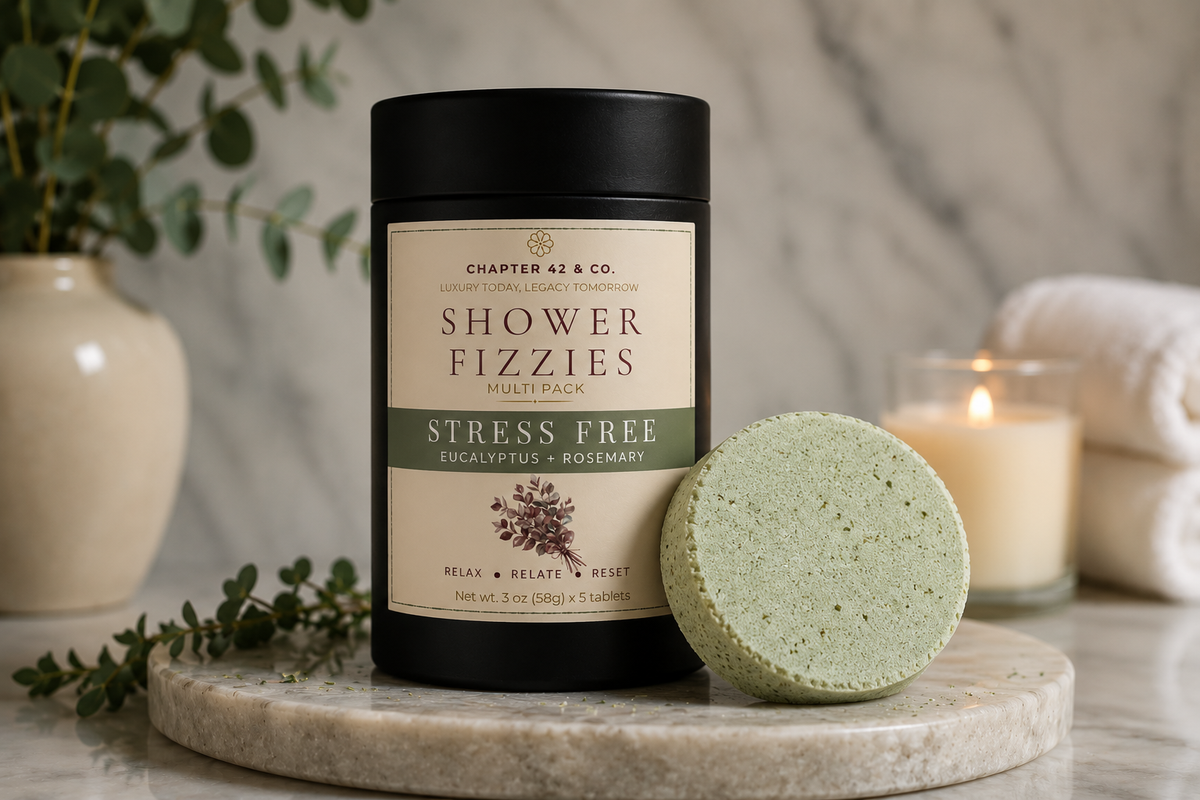 A black cylindrical container labeled 'Chapter 42 & Co. Shower Fizzies Stress Free' stands on a round stone tray beside a pale green bath bomb, with a lit candle, folded towels, and eucalyptus in the background.