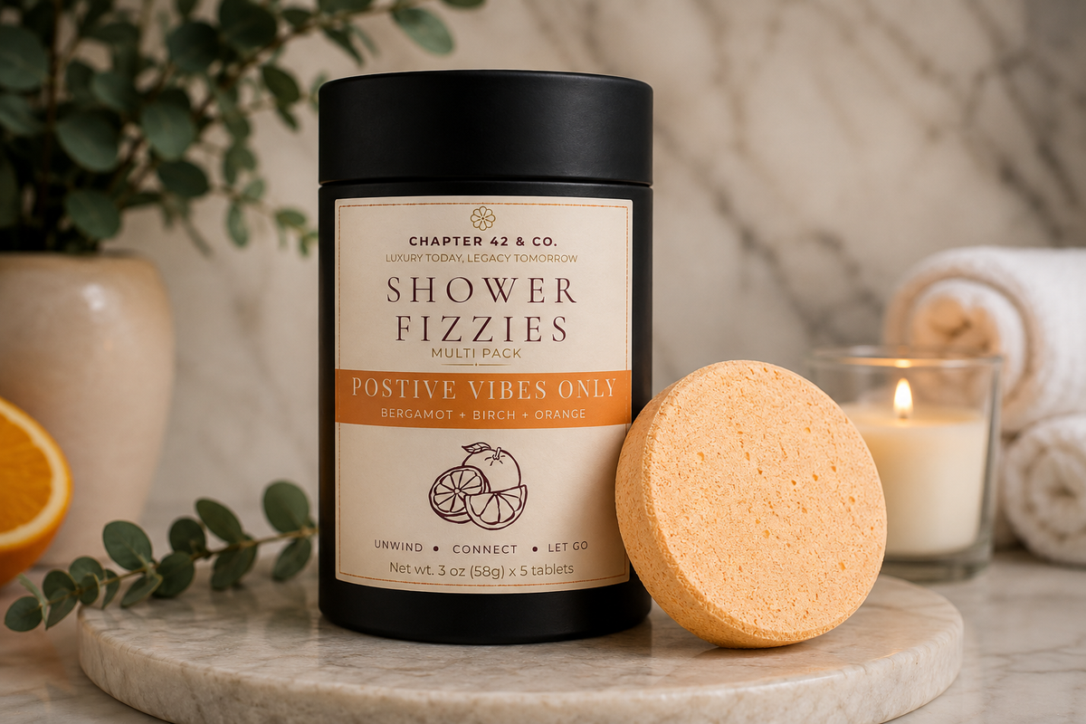 A black cylindrical jar labeled 'SHOWER FIZZIES' with a cream and orange label stands on a marble tray beside a round orange shower steamer. Rolled towels, a lit candle, and greenery are softly blurred in the background.