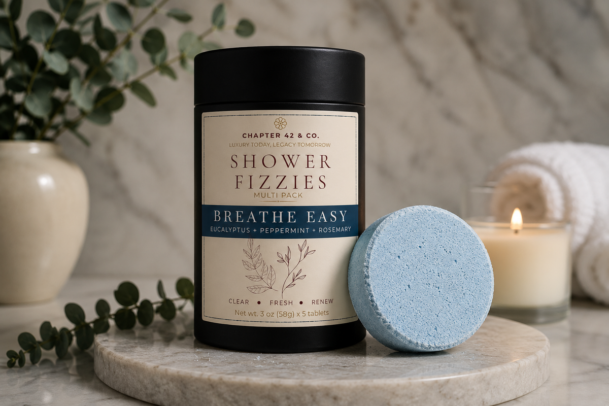 A black cylindrical container labeled 'Chapter 42 & Co. Shower Fizzies Breathe Easy' stands on a marble slab next to a round light blue shower steamer, with a lit candle, rolled towel, and greenery in the background.